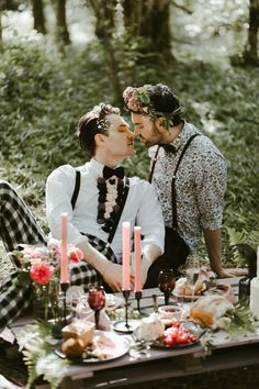 two men are sitting at a table with candles in front of them and one is kissing the other