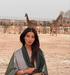 a woman standing in front of two giraffes on a dirt field with her arms crossed