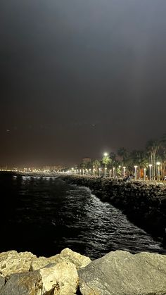 a dark sky over the ocean and beach at night with lights shining on rocks in the foreground