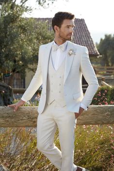 a man wearing a white suit and bow tie standing next to a wooden fence with flowers in the background