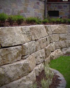 a stone wall in front of a building with green grass and bushes on the side