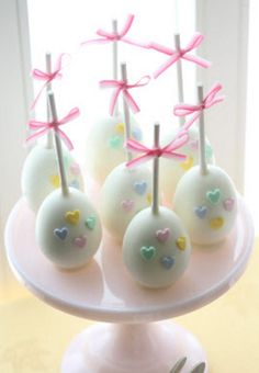 cake pops with hearts and bows on a white plate next to a cup full of silverware