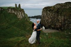 a man and woman standing on top of a lush green hillside next to the ocean