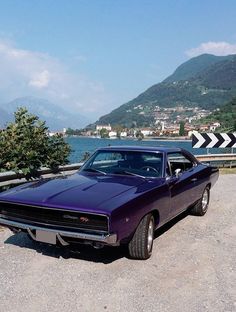 a purple muscle car parked in front of a body of water with mountains in the background