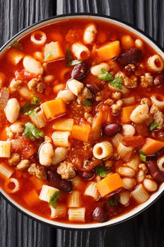 a bowl filled with pasta and bean soup on top of a wooden table next to a spoon