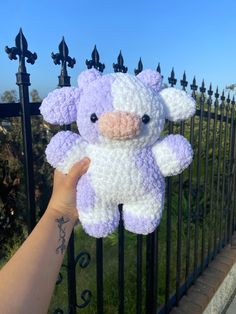 a hand holding up a purple and white stuffed animal on a black fence with green grass in the background