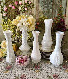 several white vases sitting on top of a table with flowers in the background and a plate next to them