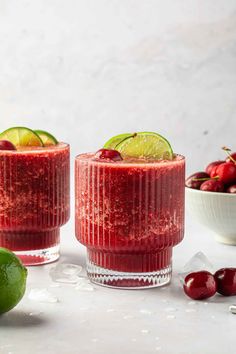 two glasses filled with cranberry smoothie next to cherries and limes