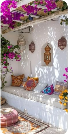 an outdoor seating area with purple flowers and hanging lanterns on the wall, in front of a white stucco building