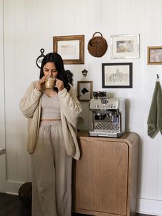 a woman standing in front of a counter drinking from a coffee cup while holding a mug