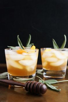 two glasses filled with ice and orange slices on top of a wooden table next to an olive branch