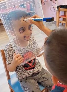 a little boy sitting in a chair with a fake face on his head and an adult brushing it's hair