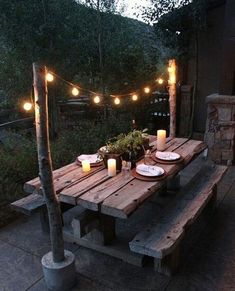 a wooden table with candles and plates on it in front of some lights hanging from the trees