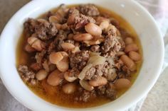 a white bowl filled with meat and beans on top of a cloth covered tablecloth