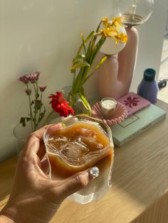 a person holding a glass filled with liquid on top of a wooden table next to flowers