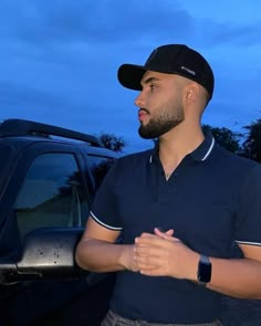 a man standing next to a car wearing a black hat and blue shirt with his hands folded