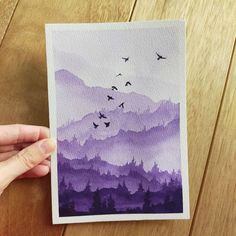 a person holding up a piece of paper with watercolor paint on it and birds flying over the mountains