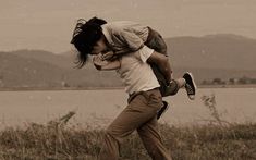 a man carrying a woman on his back while walking across a field with mountains in the background