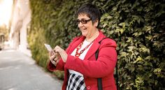 an older woman is looking at her cell phone while standing on the sidewalk in front of a green hedge