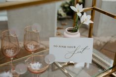 some wine glasses and a sign on a glass table with white flowers in the background