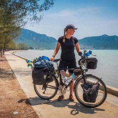 a woman is standing next to her bike on the side of the road by the water