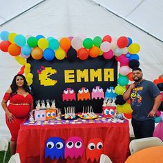 a man and woman standing in front of a table covered with balloons, pac - man cakes and other decorations