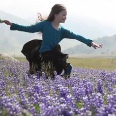 a woman riding on the back of a black horse in a field of purple flowers