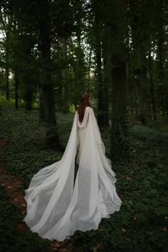 a woman in a white wedding dress is walking through the woods with her long veil over her head