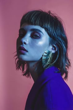 a woman with dark hair and blue eyes wearing large gold hoop earrings, standing in front of a pink background