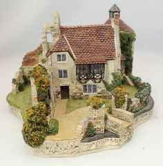 a miniature house is shown on a white surface with flowers and bushes around it,