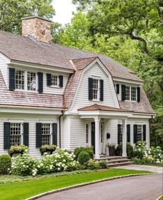 a white house with black shutters on the front and side windows, surrounded by lush green trees