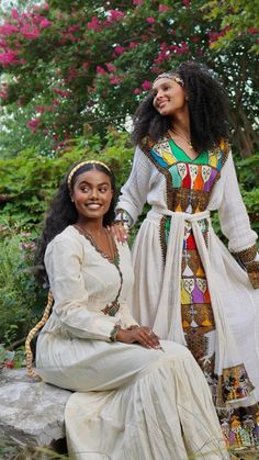 two women dressed in african clothing posing for the camera with trees and flowers behind them