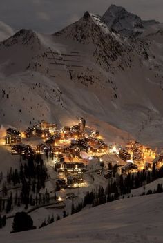 a town is lit up at night in the mountains with snow on the ground and trees around it