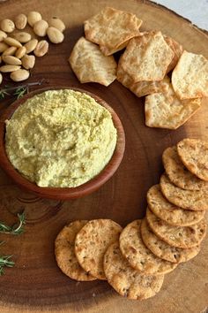 crackers and dip in a bowl on a wooden platter surrounded by nuts, herbs and other foodstuffs