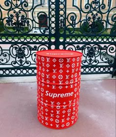 a red supreme trash can sitting in front of a wrought iron fence