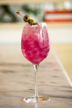 a pink drink with an olive on the top in a wine glass sitting on a counter