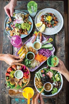 several people are eating food from bowls and plates on a wooden table with two orange juices