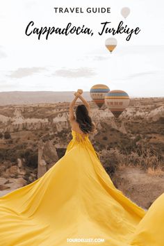 a woman in a yellow dress standing on top of a hill with hot air balloons