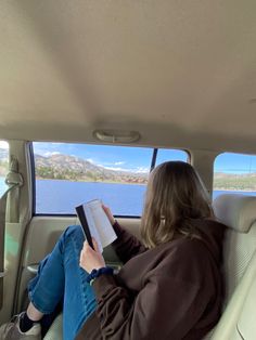 a woman sitting in the back seat of a car reading a book while looking out at water