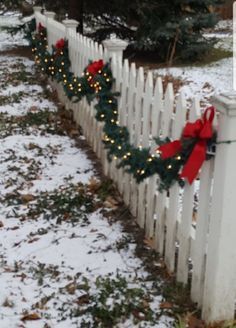 a white picket fence decorated with christmas lights and wreaths in the snow next to a pine tree