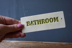 a person holding up a white tag with the word bathroom in green lettering on it