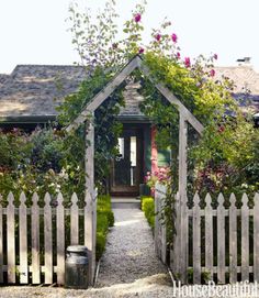 a white picket fence with flowers growing over it and a house in the background,