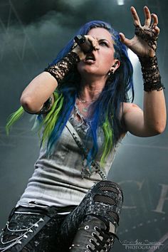 a woman with blue and green hair on stage holding her hands up in the air