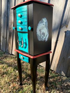 an old dresser painted black, blue and red with designs on the drawers is sitting in front of a wooden fence