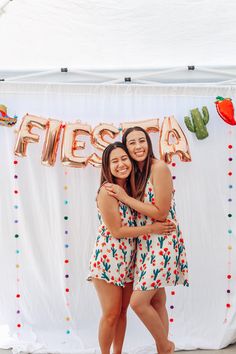 two women hugging each other in front of a sign that says fiesta with balloons and confetti