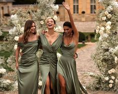 three women in long dresses are standing under an arch with white flowers and greenery