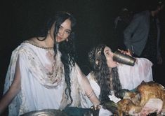 two women dressed in ancient greek garb sitting at a table with food and drinks