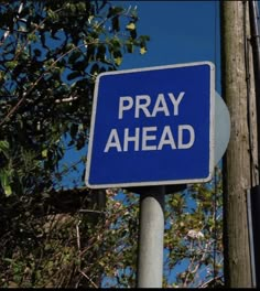 a blue and white sign that says pray ahead next to a telephone pole with trees in the background