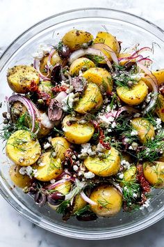 a glass bowl filled with potatoes, onions and feta cheese on top of a marble table