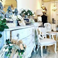 a white dresser sitting in front of a mirror next to two chairs and a table with flowers on it